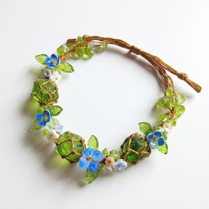 May include: A handmade bracelet featuring green glass beads encased in brown netting, accented with blue and white floral embellishments and green leaf details. The bracelet is strung on a brown cord.