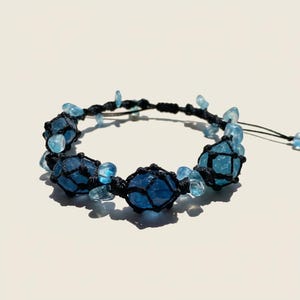 May include: A handmade bracelet featuring large, faceted blue gemstones encased in black macrame netting. Smaller blue stones are interspersed along the black cord. The bracelet is displayed on a white surface.