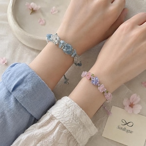 May include: Two bracelets featuring wire-wrapped beads. One bracelet displays light blue and clear faceted stones, while the other showcases pink and purple floral-shaped beads. Both pieces include small metallic accents and adjustable cord closures.