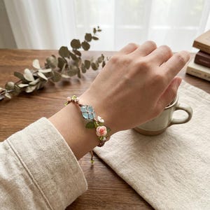 May include: A handmade bracelet featuring a light blue gemstone and delicate pink and white floral accents. The bracelet is strung on a brown cord and is adorned with green leaves. The design is set against a neutral background.