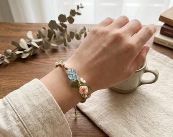 May include: A handmade bracelet featuring a light blue gemstone and delicate pink and white floral accents. The bracelet is strung on a brown cord and is adorned with green leaves. The design is set against a neutral background.