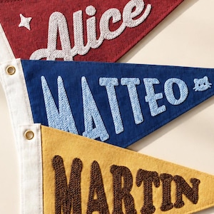 May include: Four triangular fabric pennants with white canvas headers and brass grommets. The top is navy with white embroidered text MATTEO and stars. The second is red with white text Alice and a star. The third is blue with light blue text MATTEO and a cat icon. The bottom is yellow with brown text MARTIN.