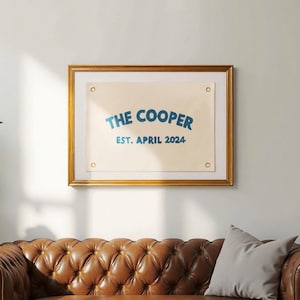 K&ouml;nnte beinhalten: Ein rechteckiges, cremefarbenes Banner aus Segeltuch mit vier Messing&ouml;sen an den Ecken, gerahmt in einem goldfarbenen Rahmen. Das Banner zeigt blauen Text mit der Aufschrift THE COOPER in gebogener Schrift und EST. APRIL 2024 darunter.