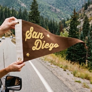Puede incluir: Un banderín triangular de fieltro marrón con el texto San Diego bordado en cursiva amarilla. El banderín tiene una franja de lona blanca en el lado izquierdo con dos ojales de latón para colgar y pequeños detalles de estrellas bordadas.
