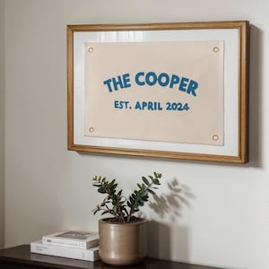 Puede incluir: Un letrero de lona enmarcado con el texto "THE COOPER EST. ABRIL 2024" en letras azules sobre un fondo crema. El marco es de madera y tiene un acabado marrón claro.