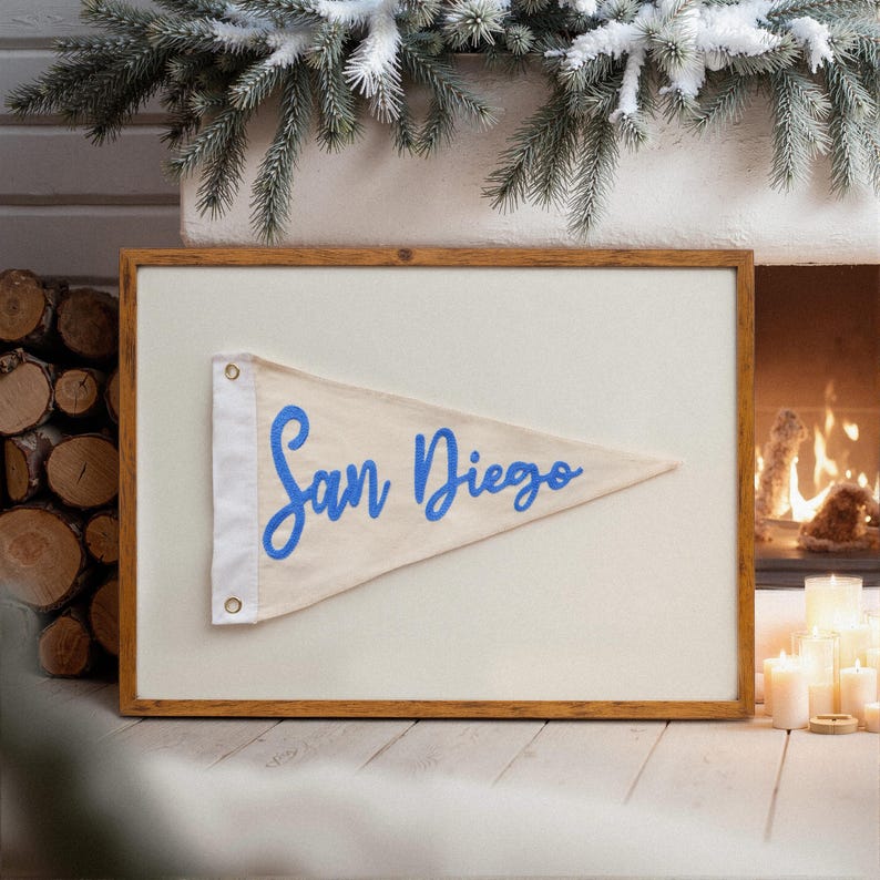 May include: A framed pennant flag with the words "San Diego" in blue script. The pennant is cream-colored and mounted on a white background within a wooden frame. The image is set against a backdrop of a fireplace and decorative greenery.