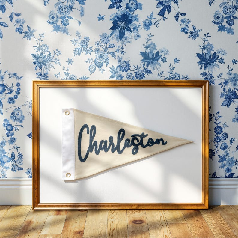 May include: A framed pennant with the word "Charleston" in navy blue script. The pennant is beige with a white border and grommets. The frame is gold, and the background is a floral wallpaper in white and blue.