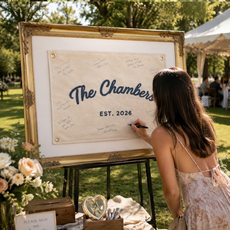 K&ouml;nnte beinhalten: Ein gro&szlig;er, rechteckiger, verzierter Goldrahmen mit einem beigen Leinwand-G&auml;stebuchschild. Die Leinwand zeigt den dunkelblauen Text 'The Chambers' und 'EST. 2026' mit handschriftlichen Unterschriften und Nachrichten um den Schriftzug.