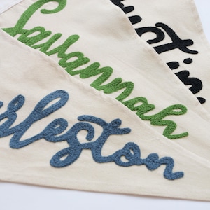 May include: Close-up of three off-white pennant flags with embroidered names in green, black, and blue. The names are "Savannah", "Austin", and "Charleston". The embroidery has a textured, raised appearance.