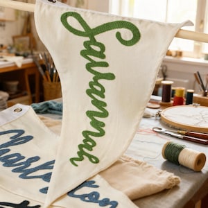 May include: Cream-colored pennant flag with the name "Lauren" embroidered in green script. The flag is hanging from a wooden dowel. Other flags with names are visible in the background, along with spools of thread and embroidery tools.