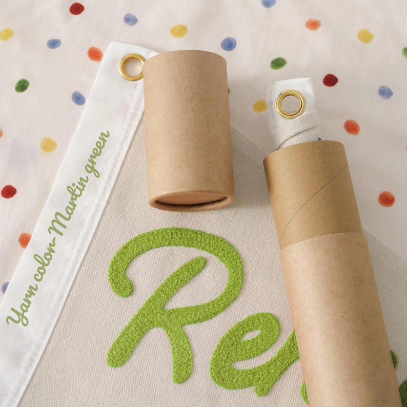 May include: A banner with the word "Re" in green, textured lettering. The banner is cream-colored with a white border that reads "Yarn color Martin green." Two brown cardboard tubes are in the image, one open and one closed. The background is white with colorful polka dots.