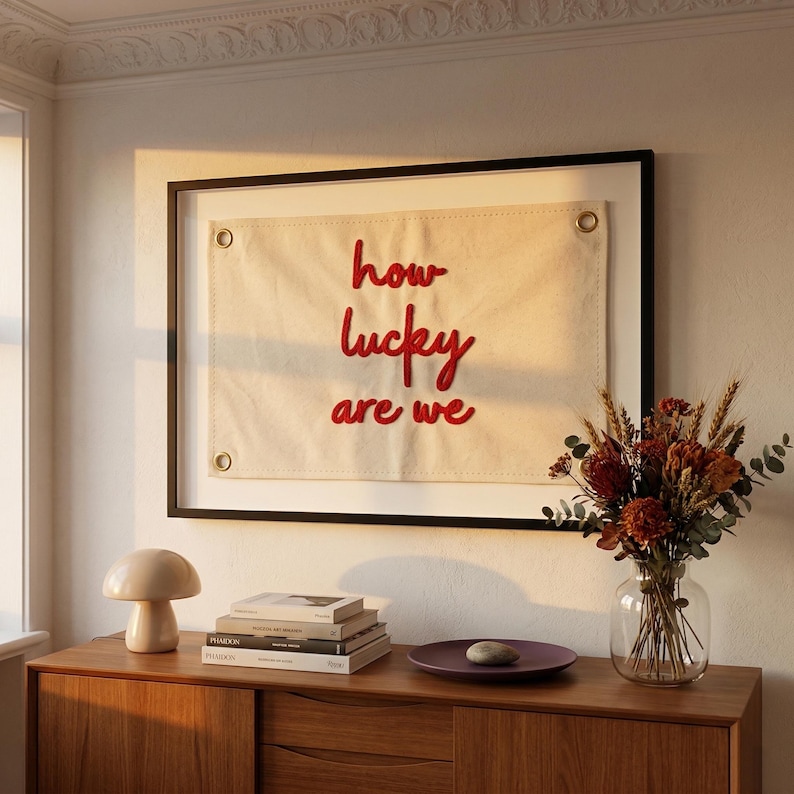 May include: A framed canvas banner with the text "how lucky are we" in red script. The banner is cream-colored and has metal grommets. The banner is displayed above a wooden cabinet with a lamp, books, and a vase of dried flowers.