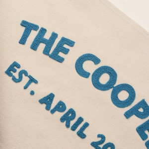K&ouml;nnte beinhalten: Eine cremefarbene Stoffoberfl&auml;che mit strukturierter, erhabener blauer Stickerei. Der Text lautet THE COOPER EST. APRIL 20, wobei die Buchstaben und Zahlen in einer fetten serifenlosen Schrift gestickt sind, was eine haptische Optik erzeugt.
