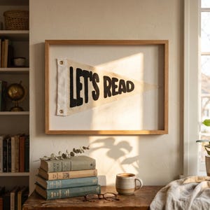 Puede incluir: Un banderín enmarcado con las palabras "LET'S READ" en letras negras. El banderín es blanco con un marco de madera. Una pila de libros, una taza y gafas están sobre una mesa de madera. Una estantería está al fondo.