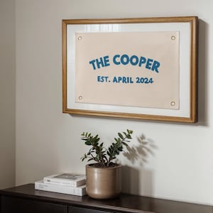 K&ouml;nnte beinhalten: Ein rechteckiges, cremefarbenes Canvas-Banner mit vier messingfarbenen Metall&ouml;sen an den Ecken, gerahmt in Holz. Das Banner zeigt blauen Text mit der Aufschrift THE COOPER in gebogener Schrift und EST. APRIL 2024 in einer geraden Zeile darunter.