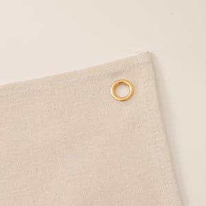 K&ouml;nnte beinhalten: Eine Ecke aus naturfarbenem, cremewei&szlig;em Segeltuch mit einer einzelnen runden, goldfarbenen Metall&ouml;se. Der Stoff weist eine sichtbare Webstruktur mit einem gen&auml;hten Saum entlang der Kante auf, was ein schlichtes Konstruktionsdetail darstellt.