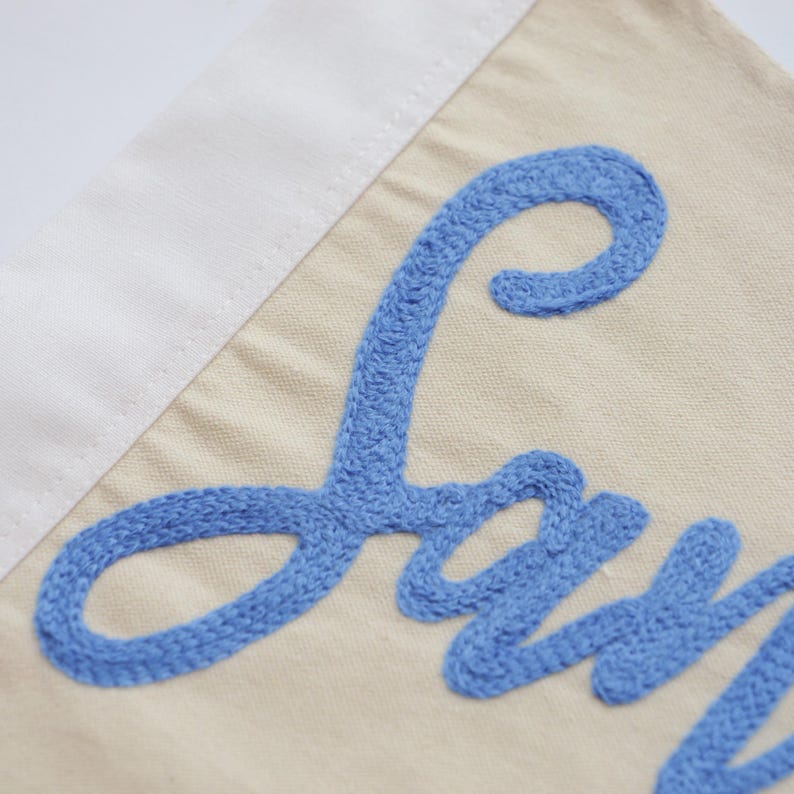 May include: Close-up of a beige fabric banner with a white border and blue embroidered text. The blue text is in a cursive font. The banner is likely a decorative item.