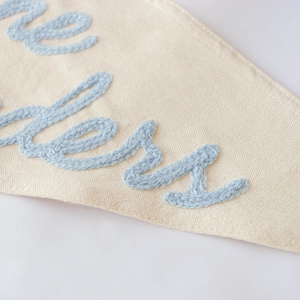 May include: Cream-colored pennant flag with light blue embroidered text. The fabric has a textured appearance. The embroidered letters are in a cursive style. The pennant is triangular in shape.