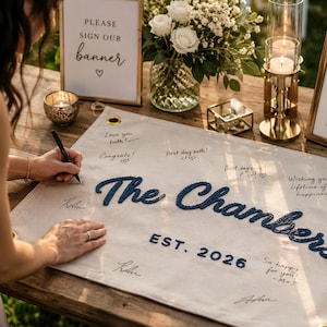 K&ouml;nnte beinhalten: Ein rechteckiges Banner aus cremefarbenem Stoff mit dem dunkelblau gestickten Text The Chambers und EST. 2026. Das Banner hat goldene Metall&ouml;sen an den Ecken und ist mit handschriftlichen Unterschriften und Nachrichten in schwarzer Tinte versehen.
