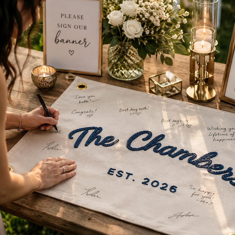 K&ouml;nnte beinhalten: Ein rechteckiges Banner aus cremefarbenem Stoff mit dem dunkelblau gestickten Text The Chambers und EST. 2026. Das Banner hat goldene Metall&ouml;sen an den Ecken und ist mit handschriftlichen Unterschriften und Nachrichten in schwarzer Tinte versehen.