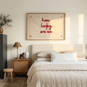May include: A bedroom scene featuring a bed with a beige quilted comforter and pillows. A framed artwork with the text "how lucky are we" in red hangs above the bed. A wooden nightstand with a lamp and decorative horse figurine is next to the bed.