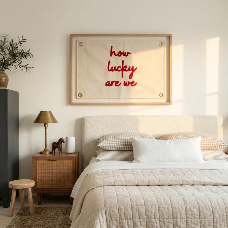 May include: A bedroom scene featuring a bed with a beige quilted comforter and pillows. A framed artwork with the text "how lucky are we" in red hangs above the bed. A wooden nightstand with a lamp and decorative horse figurine is next to the bed.