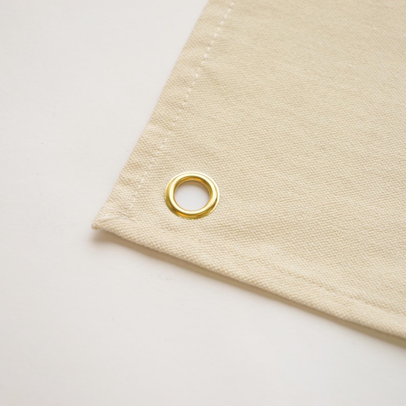 May include: Close-up of a section of beige canvas fabric with a gold-colored grommet. The fabric has a visible weave and is neatly stitched along the edges. The grommet is circular and shiny.