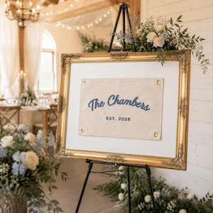 K&ouml;nnte beinhalten: Ein rechteckiges Schild mit einem cremefarbenen Stoffbanner und blau gesticktem Text The Chambers EST. 2026, befestigt mit goldenen &Ouml;sen in einem verzierten Goldrahmen auf einer schwarzen Metallstaffelei mit Blumenarrangements.