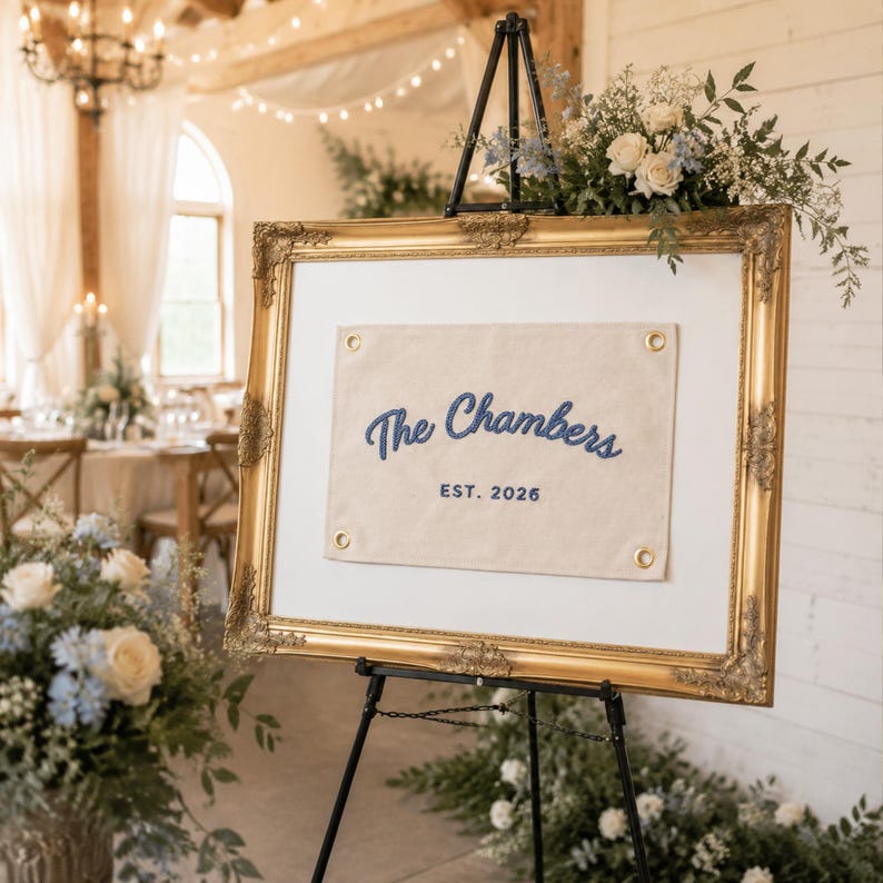 K&ouml;nnte beinhalten: Ein rechteckiges Schild mit einem cremefarbenen Stoffbanner und blau gesticktem Text The Chambers EST. 2026, befestigt mit goldenen &Ouml;sen in einem verzierten Goldrahmen auf einer schwarzen Metallstaffelei mit Blumenarrangements.