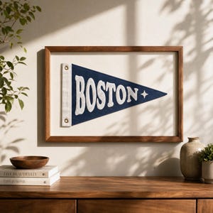 Op de afbeelding: Een ingelijste marineblauwe viltwimpel met het woord BOSTON in grote, witte, textuurrijke geborduurde letters en een klein vierpuntig sterretje aan het einde, geplaatst in een rechthoekige houten lijst met een witte achtergrond.