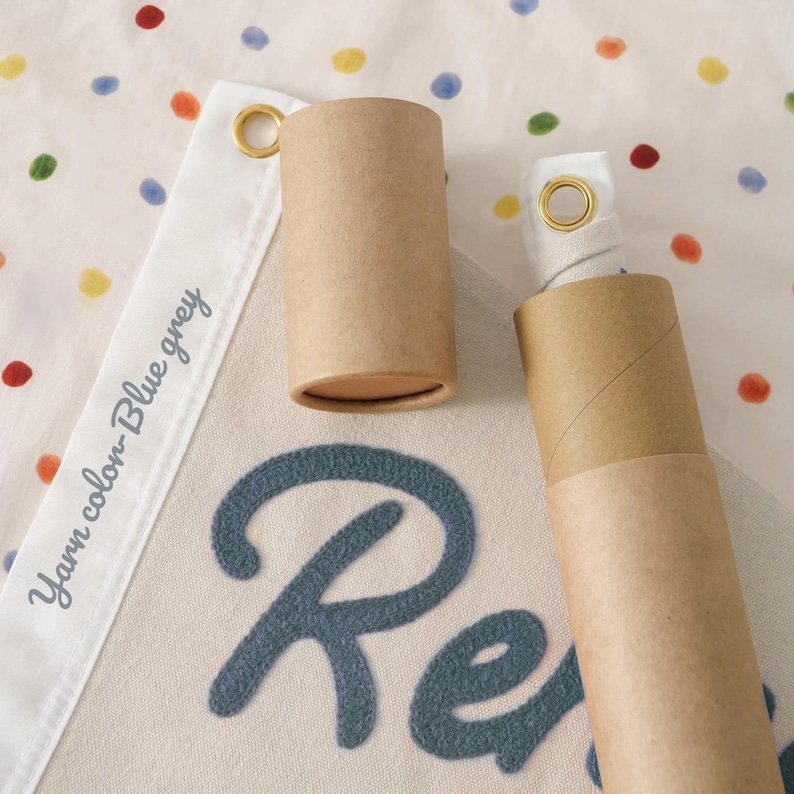 May include: A banner with the word "Re" in blue felt letters. The banner has a white border with the text "Yarn color - Blue grey". Two brown cardboard tubes are next to the banner, one open and one closed. The background is a white fabric with colorful polka dots.