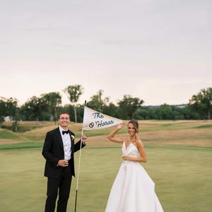 May include: A couple stands on a golf course, the man in a black tuxedo and the woman in a white wedding dress. They hold a golf flag that reads "The O'Haras" in blue script. The scene is set outdoors with green grass and trees.