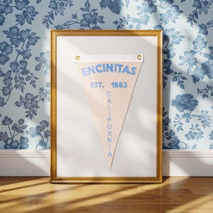Könnte beinhalten: Ein gerahmtes dreieckiges Wimpel mit dem Text "ENCINITAS EST. 1883 CALIFORNIA" in blauer Schrift auf beigefarbenem Hintergrund.