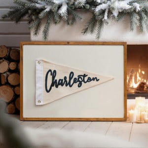 May include: A framed pennant with the word "Charleston" in navy blue script. The pennant is cream-colored and triangular, with a white border and grommets. The frame is made of wood. The background includes a fireplace and greenery.