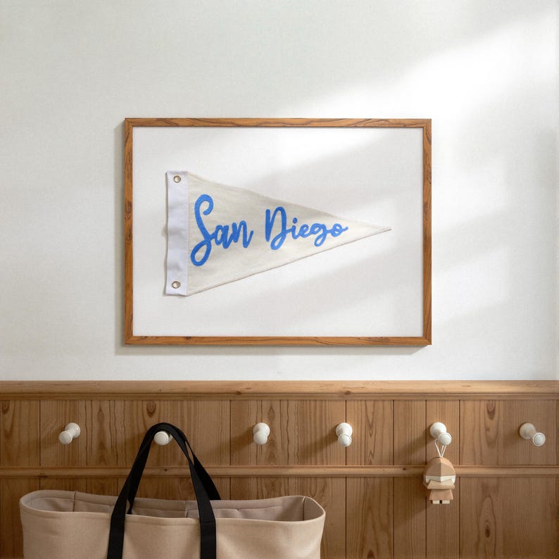 May include: A framed pennant with the words "San Diego" in blue script. The pennant is white and triangular, with a wooden frame. The image is hanging on a white wall above a wooden panel with hooks.