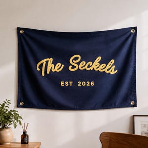 Peut inclure: Une bannière murale rectangulaire en tissu bleu marine avec le texte brodé en jaune The Seckels et EST. 2026. La bannière possède quatre œillets métalliques de couleur dorée, un à chaque coin, pour la suspendre. Le motif est centré sur le tissu.