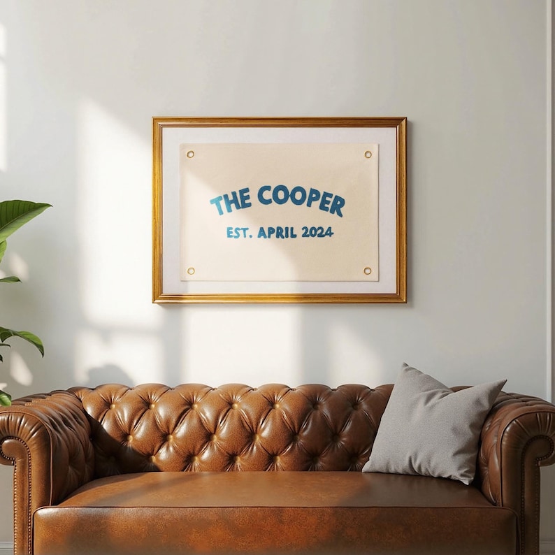 K&ouml;nnte beinhalten: Ein rechteckiges, cremefarbenes Banner aus Segeltuch mit vier Messing&ouml;sen an den Ecken, gerahmt in einem goldfarbenen Rahmen. Das Banner zeigt blauen Text mit der Aufschrift THE COOPER in gebogener Schrift und EST. APRIL 2024 darunter.