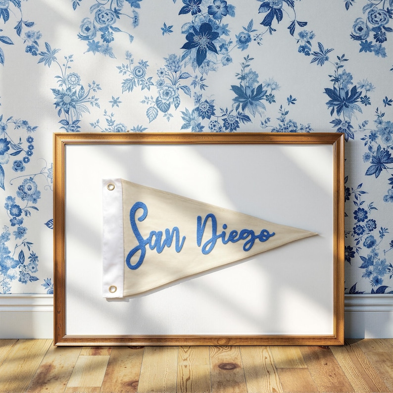 May include: A framed pennant with the text "San Diego" in blue script. The pennant is a light tan color with a white border and is mounted on a white background within a gold-colored frame. The background features a blue floral wallpaper.