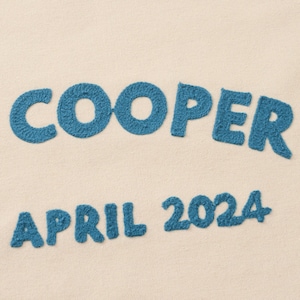 K&ouml;nnte beinhalten: Eine hellbeige Stoffoberfl&auml;che mit dunkelblauer Stickerei. Der Text lautet COOPER in einer gro&szlig;en, gebogenen Schriftart, wobei der kleinere Text APRIL 2024 direkt darunter in einer geraden Linie angeordnet ist, beides aus strukturiertem Garn.