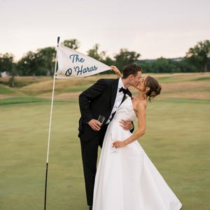May include: A newly married couple shares a kiss on a golf course. The bride wears a white wedding dress, and the groom is in a black tuxedo. A flag with the text "The O'Haras" is in the foreground.
