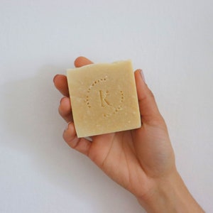 Puede incluir: Una mano sosteniendo una barra de jabón natural con un logotipo circular que dice "K" en el centro.