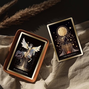 Pode incluir: Duas cartas de tar&ocirc; com detalhes dourados e fundos pretos s&atilde;o exibidas. Uma carta, rotulada "Knight of Cups", apresenta uma figura alada a cavalo. A outra carta, "The Magician", retrata uma silhueta com uma lua e estrelas.