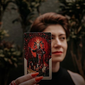 Può includere: Una carta dei tarocchi con un'illustrazione rossa e nera di una figura con corna e una corona. La carta è etichettata "XV." e "Il Diavolo."