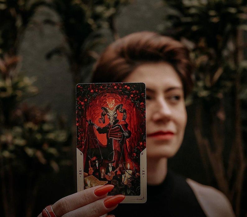 May include: A tarot card with a red and black illustration of a figure with horns and a crown. The card is labeled "XV." and "The Devil."