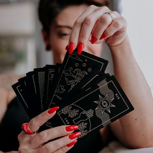 May include: A person is holding a deck of black tarot cards with gold accents. The cards are fanned out, revealing the "The Sun" card, which features a sun with a face and rays, and the "The Fool" card, which features a jester with a bell and a staff. The person's nails are painted red.