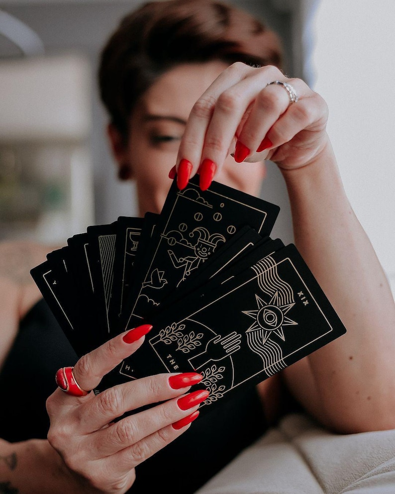 May include: A person is holding a deck of black tarot cards with gold accents. The cards are fanned out, revealing the "The Sun" card, which features a sun with a face and rays, and the "The Fool" card, which features a jester with a bell and a staff. The person's nails are painted red.