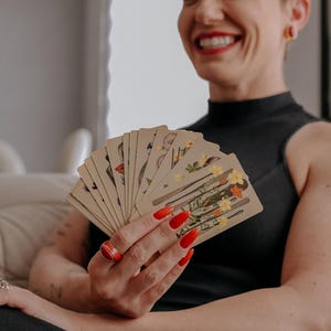 May include: A person with short, dark hair smiles while holding a fan of tarot cards with floral illustrations. The cards are fanned out in their hand, revealing the colorful images on each card.