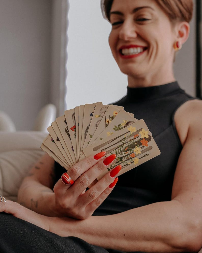 May include: A person with short, dark hair smiles while holding a fan of tarot cards with floral illustrations. The cards are fanned out in their hand, revealing the colorful images on each card.