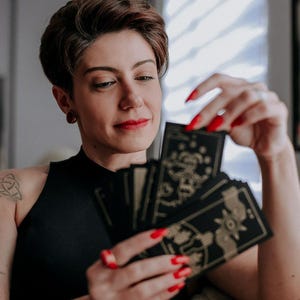 May include: A person with short brown hair is holding a spread of tarot cards with gold and black designs. The cards are fanned out in their hand, and the person is looking at the cards.