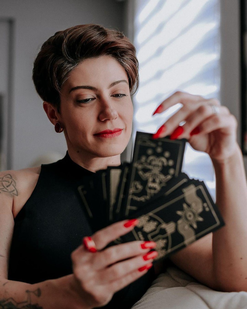 May include: A person with short brown hair is holding a spread of tarot cards with gold and black designs. The cards are fanned out in their hand, and the person is looking at the cards.